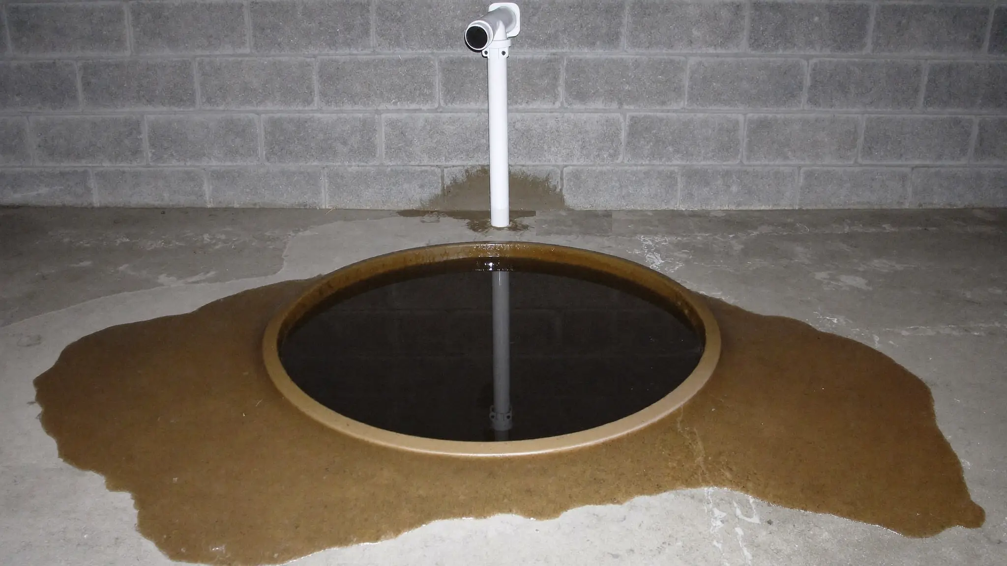 An overflowing sump pit in a basement floor with water spreading across the concrete around it